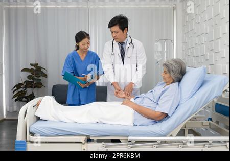 Seniorin und Ärztin. Ein asiatischer Arzt mit Stethoskop überprüft die Esel des älteren Patienten, während die junge Krankenschwester das Ergebnis in die Dokumentenmappe schreibt. Stockfoto
