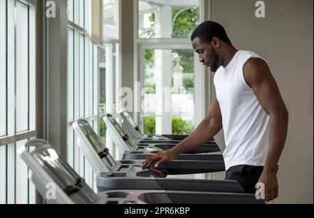 Junger, kurzer, lockig schwarzer Haarmann mit Schnurrbart und Bart, der das Laufprogramm auf dem Laufband im Fitnessstudio auswählt. Stockfoto
