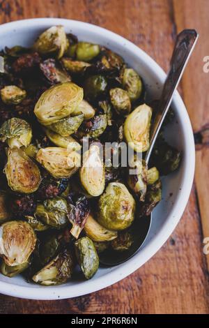 Gerösteter Rosenkohl in einer weißen Schüssel mit einem Metalllöffel auf einem braunen Holztisch. Stockfoto