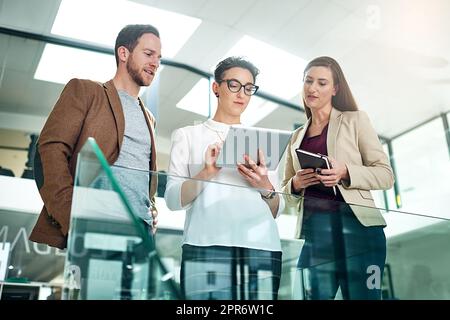 Die letzten Schliffe auf ihr Projekt zu legen. Aufnahme einer Gruppe von Kollegen, die sich über ein digitales Tablet unterhalten, während sie in einem großen, modernen Büro stehen. Stockfoto