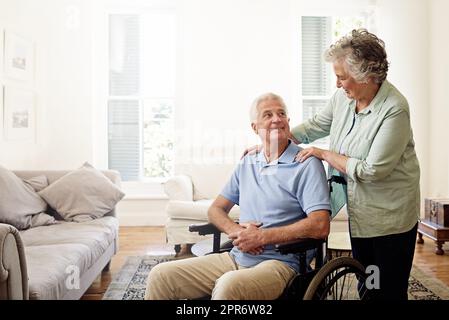 Sie sorgt immer dafür, dass sie sich wohl fühlt. Aufnahme eines lächelnden älteren Mannes im Rollstuhl und seiner Frau zu Hause. Stockfoto