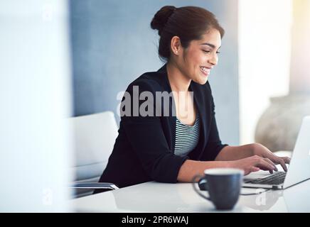 Sie arbeitet immer mit einem Lächeln im Gesicht. Eine kurze Aufnahme einer attraktiven jungen Geschäftsfrau, die in ihrem Büro an einem Laptop arbeitet. Stockfoto