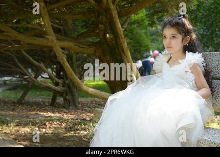 Kleines Mädchen mit einem weißen Brautkleid, das zum Geburtstag auf einer Bank sitzt Stockfoto