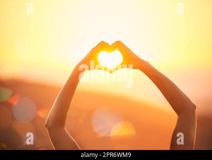 Was ist nicht zu lieben über den Sommer. Aufnahme von Händen einer nicht identifizierbaren Frau, die über einer Sonnenuntergangslandschaft eine Herzform bilden. Stockfoto