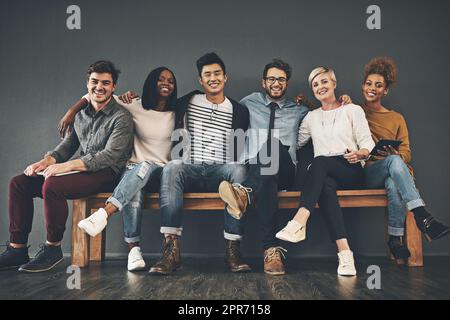 Kollegen, die Freunde geworden sind. Studioaufnahme einer vielfältigen Gruppe kreativer Mitarbeiter, die sich vor grauem Hintergrund umarmen. Stockfoto