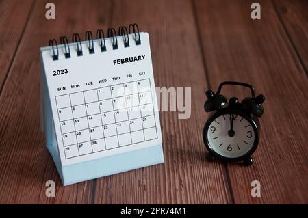 Februar 2023 weißer Tischkalender auf Holztisch mit Wecker auf 12 Uhr. Stockfoto