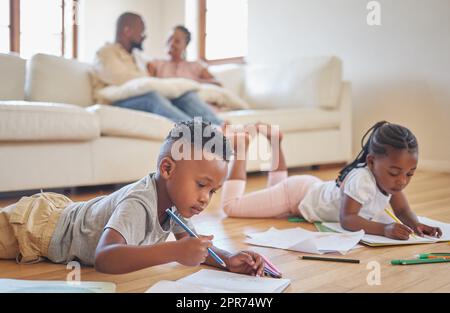 Kleine Jungen und Mädchen malen mit Malstiften auf dem Wohnzimmerboden, während sich ihre Eltern auf dem Sofa entspannen. Kleine Kinder, Schwestern und Geschwister, die sich in der Familienzeit zuhause einfärben Stockfoto