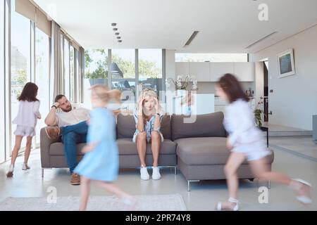 Erschöpfte Mutter und Vater, die gestresst wirkten, während Kinder in der Lounge herumlaufen und spielen. Verärgerte Eltern mit Kopfschmerzen, die auf der Couch sitzen, Kinder, die um sie herum spielen. Junge Mädchen spielen Stockfoto