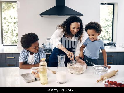 Ein süßer kleiner Junge mit Afro-Backen in der Küche zu Hause mit seiner mutter und seinem Bruder. Eine fröhliche Mischrasse, die mit Hilfe ihrer kleinen Jungs Zutaten mixt. Das Backen ist eine Bindungsaktivität Stockfoto