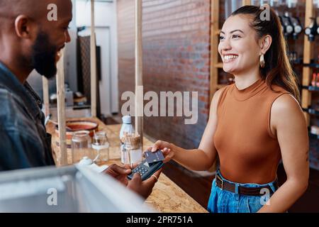 Glücklicher hispanischer Gast, der mit einer nfc-Maschine und Kreditkarte für eine Mahlzeit in einem Restaurant bezahlt. Lächelnde junge Frau, die in einem Geschäft mit ihrer Debitkarte und einem Postautomaten einkauft. Eine Frau, die die Rechnung bezahlt Stockfoto