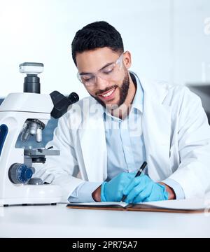 Ein hübscher junger Mann aus gemischten Rassen, der Handschuhe und einen Labormantel trägt und medizinische Proben auf einem Mikroskop in einem Labor ansieht. Ein männlicher Wissenschaftler, der eine Schutzbrille trägt und lächelt, während er in ein Notizbuch schreibt Stockfoto