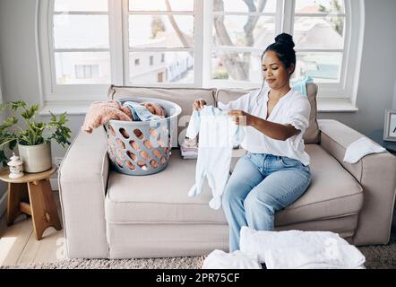 Kein einziger Fleck in Sicht. Aufnahme einer attraktiven jungen Frau, die allein in ihrem Wohnzimmer sitzt und Babykleidung zusammenfaltet. Stockfoto