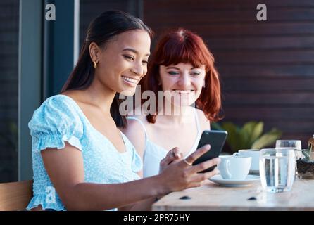 Sehen Sie sich diese neue App an. Aufnahme von zwei jungen Freundinnen, die in einem Café ein Telefon benutzen. Stockfoto