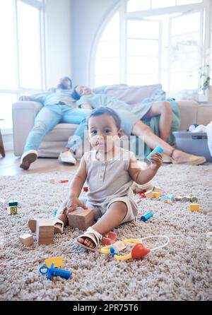 Im Handumdrehen. Aufnahme eines kleinen Mädchens, das auf dem Boden spielt, während ihre Eltern im Hintergrund schlafen. Stockfoto