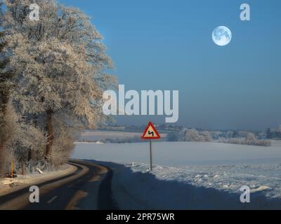 Die kurvenreiche Straße in einer ländlichen Winterlandschaft mit einem Schild am Rand der leeren Straße zur Sicherheit. Eine rutschige und nasse Straße, umgeben von Schnee an einem kalten Winterabend in der Nähe eines Waldes mit Eisfrost Stockfoto