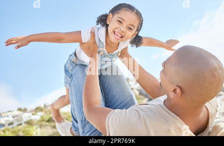 Vater und Tochter, die sich im Freien zusammentun. Porträt eines niedlichen kleinen Mädchens von unten, das Spaß hat, so zu tun, als würde es fliegen wie ein Superheld mit ausgestreckten Armen, während er von ihrem liebenden Vater gehalten wird. Stockfoto