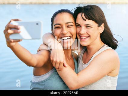 Zwei Freundinnen machen Selfies nach einem Training in der Natur und stehen mit einem Smartphone vor einem See. Junge Latino-Frau, die ihr Handy benutzt, um ein Selfie von sich und ihrer Freundin zu machen Stockfoto