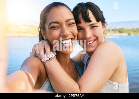 Porträt zweier lächelnder Freunde, die während der Sommerpause gemeinsam Selfies für soziale Medien auf einem See machen. Lächelnde, glückliche Frauen, die sich im Freien festklebten und umarmten Stockfoto