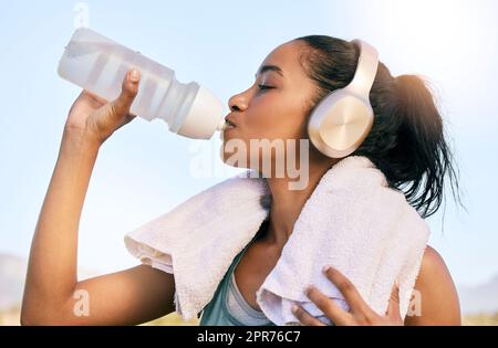 Eine Frau mit aktiver Passform, die nach dem Training im Freien kabellose Kopfhörer trägt und Wasser aus einer Flasche trinkt. Sportlerinnen löschen Durst und kühlen sich nach dem Training im Freien mit einem Handtuch ab Stockfoto