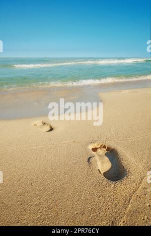 Schritte an einem Sandstrand mit einer weißen schaumigen Welle aus dem blauen Meer. Sanfte Wellen von klarem Wasser. Sandstrand mit menschlichen Fußabdrücken. Angenehmer blauer Himmel über einem friedlichen Meer. Sandiger tropischer Ozean. Stockfoto