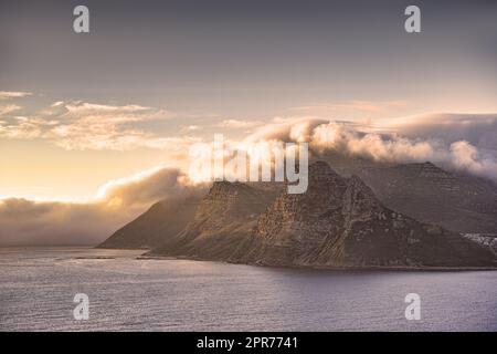 Panorama einer Bergküste mit bewölktem Sonnenuntergang in Südafrika. Malerische Landschaft mit weichen weißen Wolken, die den Tafelberg in der Dämmerung in der Nähe eines ruhigen, friedlichen Meeres an der Hout Bay in der Nähe von Kapstadt bedecken. Stockfoto