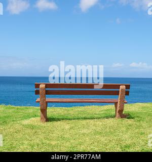 Leere Bank mit Blick auf das Meer an einem sonnigen Tag mit viel Platz. Holzsitz zum Sitzen und Reflektieren, mit therapeutischer Sicht auf Wellenmuster und Harmonie. Ein friedlicher Ort, um ruhig zu denken Stockfoto