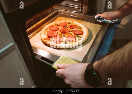 Ein Mann zieht zu Hause ein Tablett mit Peperoni-Pizza aus dem Ofen Stockfoto