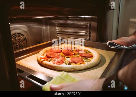 Die Hände eines Mannes ziehen eine hausgemachte Peperoni-Pizza aus dem Ofen Stockfoto