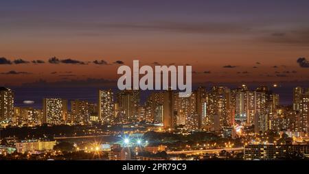 Hochhaus, Wolkenkratzer beleuchtet bei Nacht in einer Stadt mit kleinen Häusern im Vordergrund, wunderschöne moderne Gebäude und helle, leuchtende Lichter. Die blaue Skyline in der Dämmerung der Metropole. Stockfoto