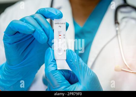 Ein asiatischer Arzt im PSA-Anzug zeigt ein positives Testergebnis mit dem Speichelantigen-Testkit zur Kontrolle des Covid-19-Coronavirus im Krankenhaus. Stockfoto