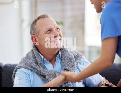 Ein älterer Mann sitzt in der Klinik, während seine Krankenschwester ihm hilft. Stockfoto