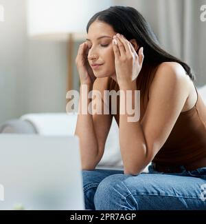 Mein Kopf tötet mich. Eine attraktive junge Frau, die allein in ihrem Wohnzimmer sitzt und sich gestresst fühlt, während sie ihren Laptop benutzt. Stockfoto
