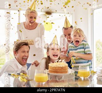 Eine fröhliche, generationsfreudige Familie trägt Partyhüte, während sie den Geburtstag kleiner Mädchen mit Konfetti und Kuchen zu Hause feiert. Kinder, Eltern und Großeltern freuen sich auf eine besondere Überraschungsveranstaltung Stockfoto