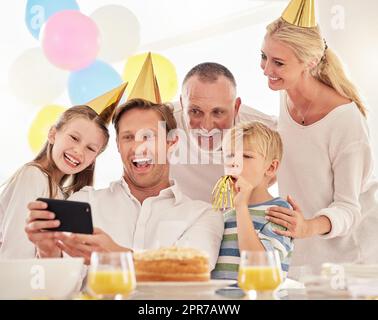 Eine glückliche Familie feiert einen Geburtstag mit einer Party, trägt Hüte und macht ein Selfie am Telefon. Reifer Mann, der auf einer Party ein Foto von Vater, Frau und Kindern macht und dabei besondere Erinnerungen weckt Stockfoto