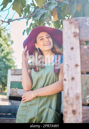 Porträt einer Frau mit Strohhut, die unter einem Baum neben einer rustikalen Holzkiste steht. Eine junge, glückliche Frau trägt einen Sommerhut und ein Latzkleid in einem Garten an einem sonnigen Tag beim Äpfelpflücken Stockfoto