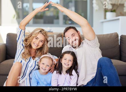 Lächelndes Paar mit kleinen Kindern, die sitzen und ein symbolisches Dach mit Kinderhänden bauen. Fröhliche Familie, die am Umzugstag im neuen Haus auf dem Boden saß. Durch Versicherung abgedeckt und geschützt Stockfoto