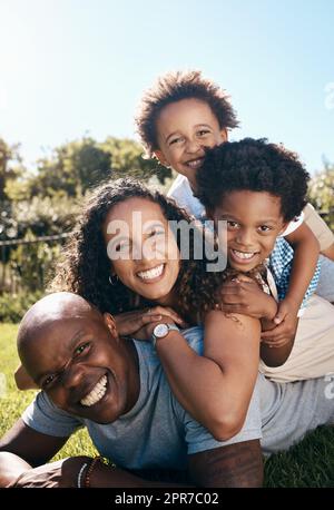 Eine glückliche afroamerikanische Familie von vier Personen, die übereinander liegen, während sie Spaß haben und zusammen in der Sonne spielen. Sorgenfreie Mutter und zwei Kinder stapelten sich auf Vater, während sie im Park eine Beziehung knüpften Stockfoto