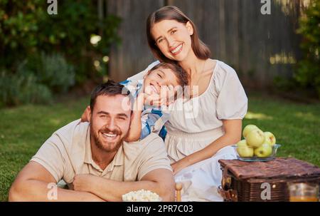 Eine glückliche Familie mit einem Kind, die ein Picknick im Garten macht. Porträt eines lächelnden, süßen Jungen mit seinen Eltern, die sich im Garten entspannen. Eine Mutter und ein Vater, die sich mit ihrem Sohn auf dem Rasen amüsieren Stockfoto