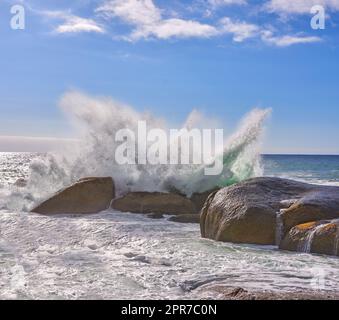 Ozeanwellen stürzen an einem felsigen Strand und Ufer unter blauem Himmel in Camps Bay, Kapstadt, Südafrika. Malerische Meereslandschaft mit Gezeitenmeer, das über Felsen und Felsbrocken auf der Touristenattraktion weht Stockfoto