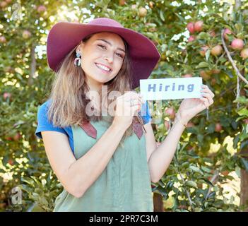 Eine Apfelbauerin, die Arbeiter anheuert, um während der Ernte auf ihrer Obstfarm zu helfen. Porträt einer glücklichen jungen Frau, die Jobs anpreist und eine Anwerbung in einem Obstgarten findet, an einem sonnigen Tag in der Nähe von Bäumen. Stockfoto