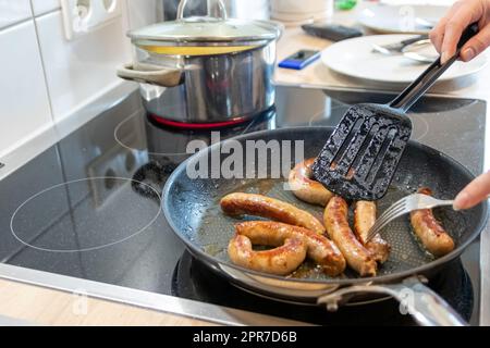 Frau, die Würstchen in der Pfanne auf Küchenplatte mit Gabel und schwarzem Spatel mit köstlicher fetter Bratwurst zum Abendessen kocht, als ungesunde, aber würzige und geröstete Wurstmahlzeit zum Mittagessen und die Kinder Stockfoto