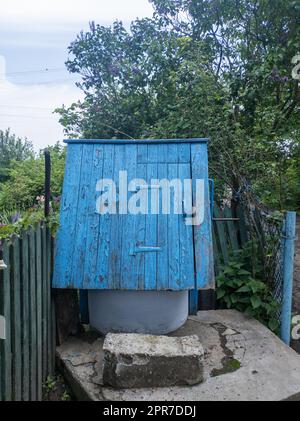 Uralter artesischer Brunnen und Holzdach mit hängendem Holzeimer im Dorf inmitten der Bäume. Stockfoto