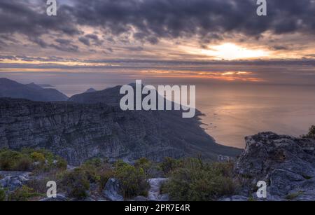 Blick über eine Bergküste bei Sonnenuntergang in Südafrika. Malerische Landschaft mit dunklen Wolken über einem ruhigen und friedlichen Ozean in der Nähe von Kapstadt mit der Sonne hinter grauen Wolken am Himmel und Kopierraum Stockfoto