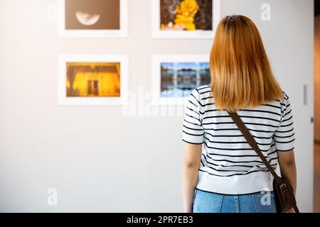 Asiatische Frau steht und schaut Kunstgalerie vor farbenfrohen gerahmten Bildern an Stockfoto