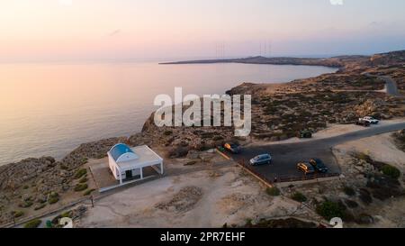 Luftaufnahme des Sonnenuntergangs an der Küste und Wahrzeichen der weißen gewaschenen Kapelle Agioi Anargyroi, Cavo Greco Protaras, Famagusta, Zypern von oben. Vogelperspektive wetteifern Stockfoto