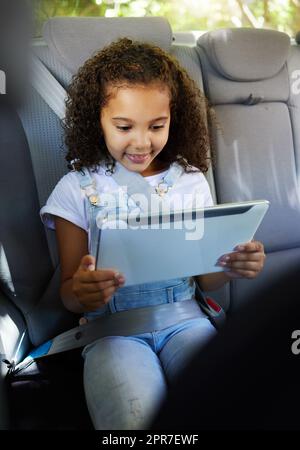 Sich auf dem Laufwerk beschäftigt zu halten. Ein kurzer Schuss eines entzückenden kleinen Mädchens, das ihr Tablet benutzt, während es auf dem Rücksitz eines Autos sitzt. Stockfoto