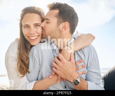 Wunderschöne Frau mittleren Alters, die von ihrem Mann auf die Wange geküsst wird, während sie draußen am Strand sitzt. Liebevolles Ehepaar, das sich bei einem romantischen Date umarmt und seiner Frau draußen Liebe zeigt Stockfoto