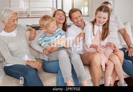 Porträt einer glücklichen Familie, die lacht und auf der Couch im Wohnzimmer sitzt. Ein Mädchen und ein Junge, die sich mit Eltern und Großeltern im Wohnzimmer anfreunden. Großeltern besuchen die Enkel Stockfoto