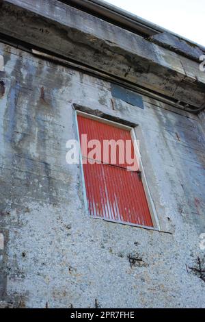 Ein Steingebäude mit einer roten Tür auf einem alten, verlassenen Grundstück. Architektonische Details des Äußeren eines grauen Industriegebäudes. Verwitterte Betonwände mit einer hohen Holztür Stockfoto