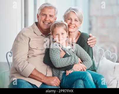 Das kleine Mädchen und ihre Großeltern schauen in die Kamera, während sie zu Hause zusammen sitzen. Süßes Mädchen, das sich mit ihrer Großmutter und ihrem Großvater verbindet. Liebevolles Paar, das Zeit mit seiner Enkelin verbringt Stockfoto